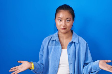 Téléchargez les photos : Asiatique jeune femme debout sur fond bleu clueless et confus avec les bras ouverts, aucune idée concept. - en image libre de droit
