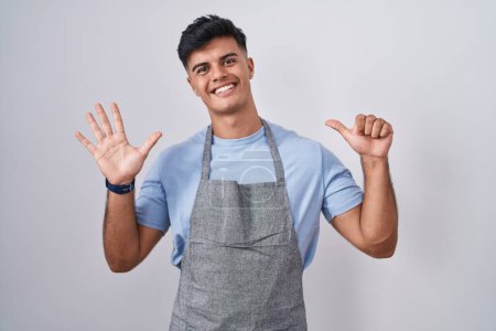 Foto de Joven hispano vistiendo delantal sobre fondo blanco mostrando y señalando con los dedos número seis mientras sonríe confiado y feliz. - Imagen libre de derechos