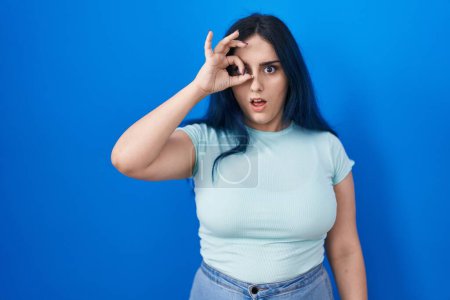 Photo for Young modern girl with blue hair standing over blue background doing ok gesture shocked with surprised face, eye looking through fingers. unbelieving expression. - Royalty Free Image