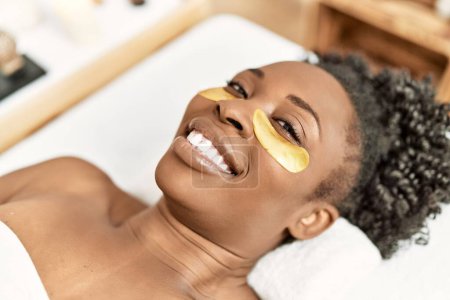 Téléchargez les photos : Femme afro-américaine allongée sur une table de massage ayant un traitement de masque oculaire au salon de beauté - en image libre de droit