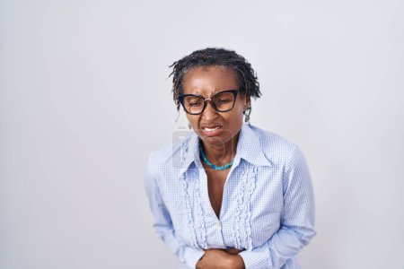 Téléchargez les photos : Femme africaine avec dreadlocks debout sur fond blanc portant des lunettes avec la main sur le ventre parce que les nausées, la maladie douloureuse se sentent mal. concept de douleur. - en image libre de droit