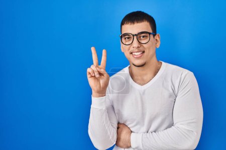 Photo for Young arab man wearing casual white shirt and glasses smiling with happy face winking at the camera doing victory sign. number two. - Royalty Free Image