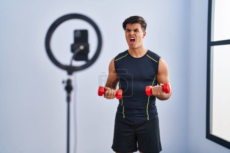 Photo for Hispanic man recording training tutorial at the gym angry and mad screaming frustrated and furious, shouting with anger. rage and aggressive concept. - Royalty Free Image