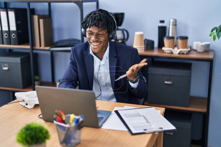 Téléchargez les photos : Jeune homme africain avec dreadlocks travaillant au bureau portant casque célébrant la réalisation avec sourire heureux et expression gagnante avec la main levée - en image libre de droit