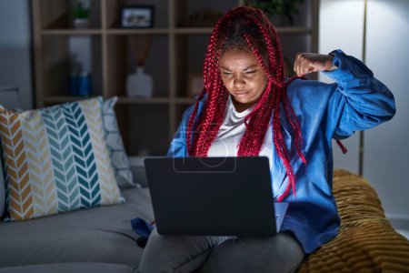 Téléchargez les photos : Femme afro-américaine avec des cheveux tressés en utilisant ordinateur portable la nuit personne forte montrant muscle du bras, confiant et fier de la puissance - en image libre de droit