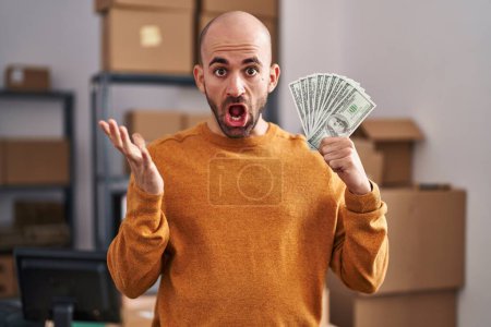 Photo for Young bald man with beard working at small business ecommerce holding money celebrating victory with happy smile and winner expression with raised hands - Royalty Free Image