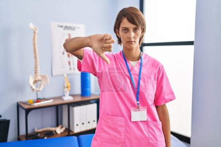 Photo for Brunette woman working at rehabilitation clinic looking unhappy and angry showing rejection and negative with thumbs down gesture. bad expression. - Royalty Free Image