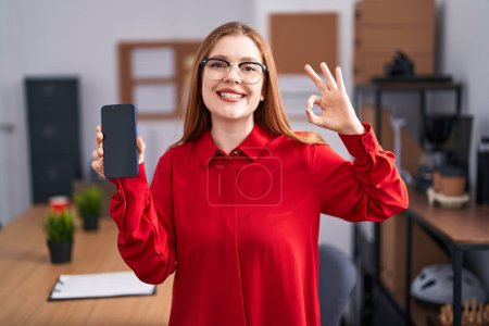 Téléchargez les photos : Femme rousse travaillant au bureau montrant l'écran du smartphone faire ok signe avec les doigts, sourire geste amical excellent symbole - en image libre de droit