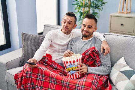 Téléchargez les photos : Couple homosexuel manger pop-corn regarder la télévision souriant regardant vers le côté et regardant loin en pensant. - en image libre de droit