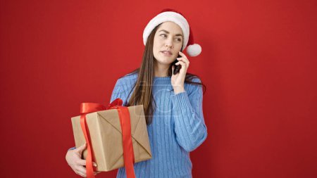 Téléchargez les photos : Jeune femme caucasienne portant chapeau de Noël parler sur smartphone tenant cadeau sur fond rouge isolé - en image libre de droit