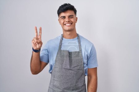 Foto de Joven hispano vistiendo delantal sobre fondo blanco mostrando y señalando con los dedos número dos mientras sonríe confiado y feliz. - Imagen libre de derechos