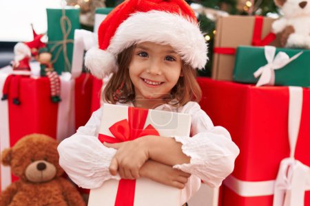 Photo for Adorable hispanic girl hugging christmas gift sitting on floor at home - Royalty Free Image