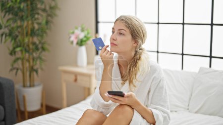 Photo for Young blonde woman wearing bathrobe shopping with credit card and smartphone thinking at bedroom - Royalty Free Image