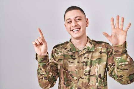 Foto de Joven vestido con camuflaje uniforme del ejército mostrando y señalando hacia arriba con los dedos número seis mientras sonríe confiado y feliz. - Imagen libre de derechos