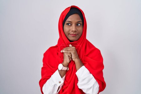 Foto de Mujer árabe joven vistiendo bufanda islámica tradicional hijab riendo nerviosa y emocionada con las manos en la barbilla mirando a un lado - Imagen libre de derechos