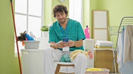Photo for Young hispanic man professional cleaner using smartphone relaxing at laundry room - Royalty Free Image