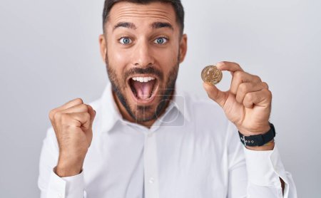 Téléchargez les photos : Bel homme hispanique tenant litecoin pièce crypto-monnaie criant fier, célébrant la victoire et le succès très excité avec les bras levés - en image libre de droit