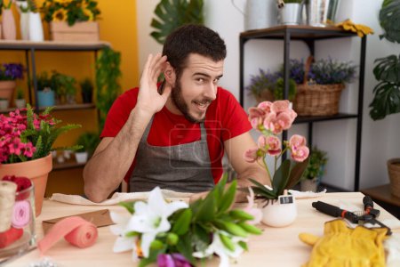 Téléchargez les photos : Jeune homme hispanique travaillant à la boutique de fleuriste souriant avec la main sur l'oreille écouter et entendre des rumeurs ou des commérages. concept de surdité. - en image libre de droit