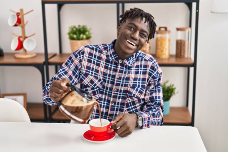Téléchargez les photos : Africain américain verser du café sur la tasse assis sur la table à la maison - en image libre de droit
