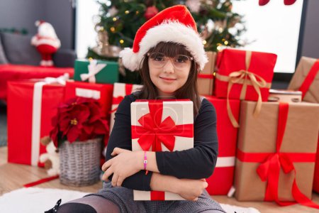 Foto de Adorable chica hispana abrazando regalo de Navidad sentada en el suelo en casa - Imagen libre de derechos