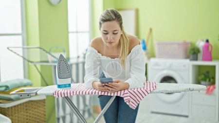 Photo for Young blonde woman using smartphone leaning on ironing table at laundry room - Royalty Free Image
