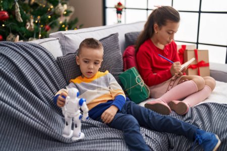 Photo for Brother and sister playing with robot and writing on notebook sitting on sofa by christmas tree at home - Royalty Free Image