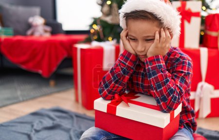 Photo for Adorable hispanic boy holding christmas gift sitting on floor with unhappy expression at home - Royalty Free Image