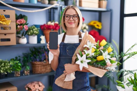 Téléchargez les photos : Jeune femme caucasienne travaillant à la boutique de fleuristes montrant écran smartphone souriant regardant vers le côté et regardant loin en pensant. - en image libre de droit