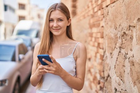 Téléchargez les photos : Jeune femme caucasienne souriant confiant en utilisant un smartphone à la rue - en image libre de droit