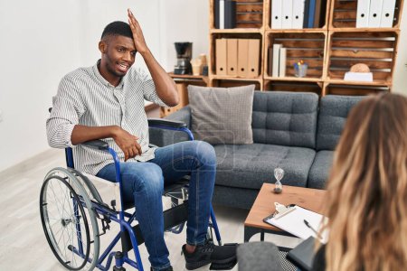 Téléchargez les photos : L'homme afro-américain faisant une thérapie assis sur un fauteuil roulant surpris avec la main sur la tête pour erreur, rappelez-vous erreur. oublié, mauvais concept de mémoire. - en image libre de droit