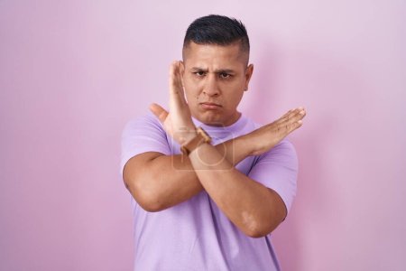Téléchargez les photos : Young hispanic man standing over pink background rejection expression crossing arms doing negative sign, angry face - en image libre de droit
