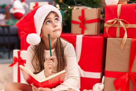 Téléchargez les photos : Adorable hispanique fille écriture santa claus lettre assis sur le sol par arbre de Noël à la maison - en image libre de droit