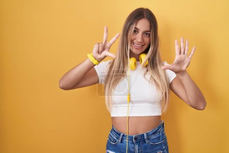 Foto de Mujer rubia joven de pie sobre fondo amarillo con auriculares que muestran y señalan hacia arriba con los dedos número ocho mientras sonríe confiado y feliz. - Imagen libre de derechos
