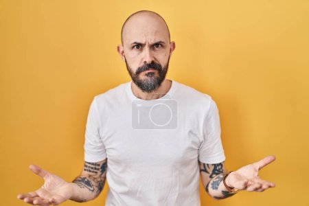 Photo for Young hispanic man with tattoos standing over yellow background clueless and confused with open arms, no idea concept. - Royalty Free Image