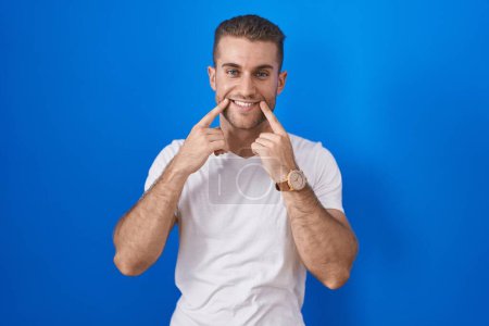Téléchargez les photos : Jeune homme caucasien debout sur fond bleu souriant avec la bouche ouverte, les doigts pointant et forçant sourire joyeux - en image libre de droit