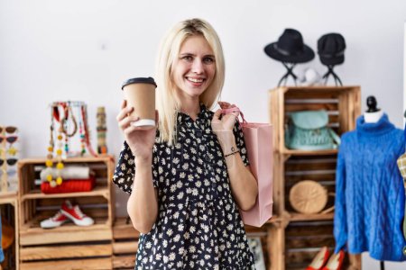 Foto de Joven mujer rubia sonriendo confiada sosteniendo bolsas de compras y café en la tienda de ropa - Imagen libre de derechos