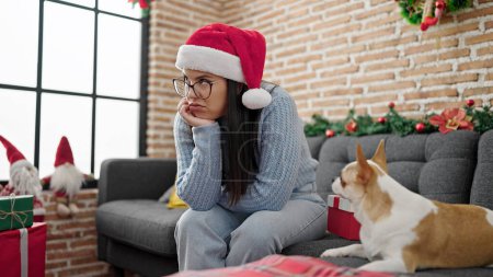 Foto de Joven mujer hispana con perro chihuahua usando sombrero de navidad desempacando regalo buscando molesto en casa - Imagen libre de derechos