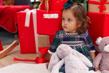 Téléchargez les photos : Adorable bambin blonde assis sur le sol par des cadeaux de Noël avec une expression détendue à la maison - en image libre de droit