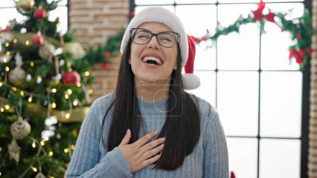Téléchargez les photos : Jeune femme hispanique riant debout près de l'arbre de Noël avec la main sur la poitrine à la maison - en image libre de droit