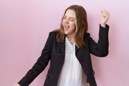 Téléchargez les photos : Jeune femme d'affaires caucasienne portant une veste noire dansant heureuse et joyeuse, souriante, décontractée et confiante, écoutant de la musique - en image libre de droit