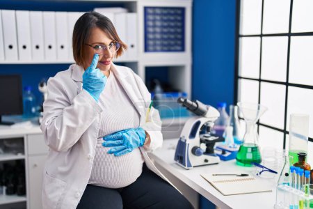 Foto de Mujer embarazada que trabaja en el laboratorio científico apuntando al ojo observándote gesto, expresión sospechosa - Imagen libre de derechos