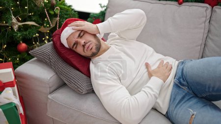 Téléchargez les photos : Jeune homme hispanique souffrant de maux d'estomac portant un chapeau de Noël à la maison - en image libre de droit