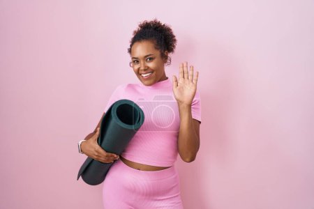 Photo for Young hispanic woman with curly hair holding yoga mat over pink background waiving saying hello happy and smiling, friendly welcome gesture - Royalty Free Image