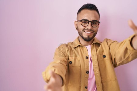 Photo for Young hispanic man standing over pink background looking at the camera smiling with open arms for hug. cheerful expression embracing happiness. - Royalty Free Image