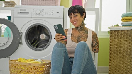 Photo for Hispanic woman with amputee arm using smartphone sitting by washing machine at laundry room - Royalty Free Image