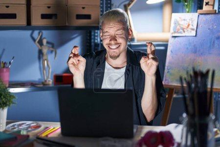 Téléchargez les photos : Jeune homme caucasien utilisant ordinateur portable la nuit au studio d'art geste doigt croisé sourire avec espoir et les yeux fermés. chance et concept superstitieux. - en image libre de droit