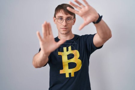 Photo for Caucasian blond man wearing bitcoin t shirt doing frame using hands palms and fingers, camera perspective - Royalty Free Image