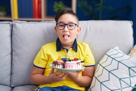 Foto de Joven niño hispano sosteniendo dulces de pastel sacando la lengua feliz con expresión divertida. - Imagen libre de derechos