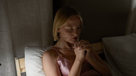 Téléchargez les photos : Jeune femme blonde assise sur le lit priant dans la chambre - en image libre de droit