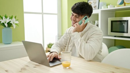 Photo for Young hispanic man using laptop talking on smartphone sitting on table at dinning room - Royalty Free Image